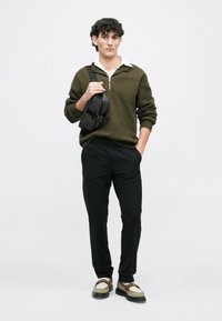 Giovane con capelli ricci che indossa un maglione verde oliva, pantaloni neri, mocassini e una borsa a tracolla nera su uno sfondo bianco.