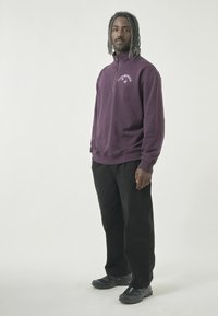 Lila Zip-Up Sweatshirt mit hohem Kragen, versehen mit einem kleinen Grafiklogo. Tragend mit schwarzen Hosen und schwarzen Sportschuhen.