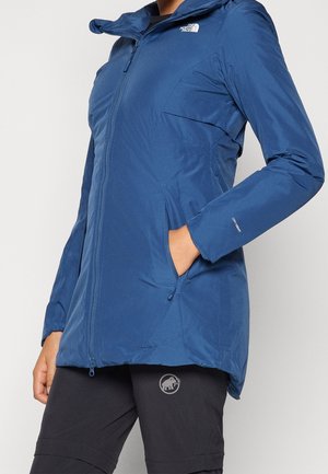 Personne portant une veste North Face bleue zippée avec les mains dans les poches, associée à un pantalon noir Mammut sur un fond uni.