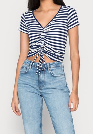 Kobieta ubrana w granatowo-biały pasiasty marszczony crop top z dekoltem w kształcie litery V, w połączeniu z jasnoniebieskimi jeansami z wysokim stanem na jednolitym tle.