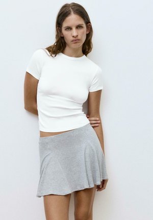 Nuori nainen valkoisessa lyhythihaisessa taâssissa ja harmaassa liehuvassa hameessa, seisoo valkoista seinää vasten, kädet selän takana.