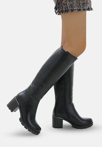 Las botas negras hasta la rodilla están hechas de un material sintético liso, cuentan con un tacón grueso, punta redondeada y una cremallera lateral para ponérselas fácilmente. Suela texturizada para mayor agarre.