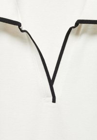 Polo blanc avec un col en V noir. Le tissu présente une texture. Le col comporte des coutures noires contrastantes.