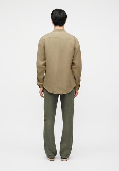 Άνδρας που στέκεται με την πλάτη, φοράει μπεζ μακρυμάνικο πουκάμισο, χακί παντελόνι και πλεκτά παπούτσια χωρίς κορδόνια μπροστά σε απλό φόντο.