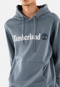 Felpa grigia in cotone con tasca a marsupio frontale, cappuccio con drawstring e il testo "Timberland" bianco con grafica del logo.