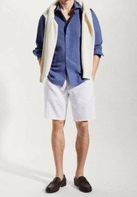 Camicia blu in lino con bottoni, pantaloni corti bianchi in lino, maglione beige in maglia appoggiato sulle spalle e mocassini marroni. Abbigliamento estivo casual.