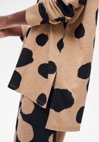 Brązowa bluzka z długim rękawem w duże czarne grochy, z luźnym krojem i rozcięciami po bokach dla wygody. Miękki materiał z widocznymi detalami szycia.