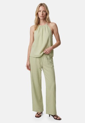 Kobieta ubrana w jasnozielony bezrękawnik z wiązaniem na szyi i pasujące szerokie spodnie oraz czarne sandały na obcasie, stojąca na jednolitym tle.