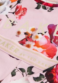 Różowa tkanina z kwiatowym wzorem z różami i stokrotkami. Tekst brzmi "BAKER BY TED BAKER" w złotym kolorze, z złotą akcentowaną wstążką.