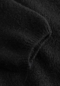 Fekete kötött anyag puha, bolyhos textúrával. Bordázott mandzsettával és változó felületi mintázattal rendelkezik, amely melegséget és vastagságot sugall.