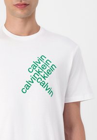 Hvid bomulds t-shirt med diagonal grøn tekst, der gentager "calvin klein," med en rund halsudskæring og korte ærmer.