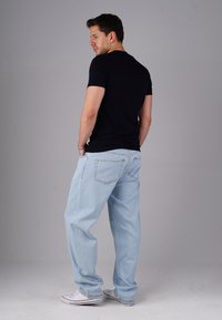 Man wearing black t-shirt, light blue jeans, and white sneakers standing with hands in pockets, looking to the side against plain background.