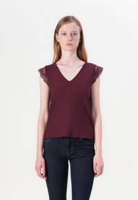 Burgundské tričko s výstřihem do V a krajkovými krátkými rukávy, vyrobené z měkké látky. Zobrazeno s tmavě modrými džíny, které mají jednoduchý, rovný střih.