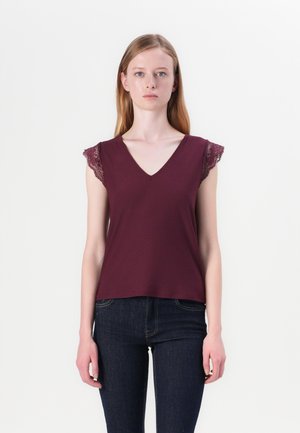 Burgunderfarget V-hals topp med blondeermet, laget av mykt stoff. Vist sammen med mørkeblå jeans, med en enkel, rett snitt.