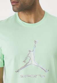 T-shirt in cotone verde menta con un logo Jumpman lucido e riflettente e il testo "SPORT" in grassetto sottostante. Collo rotondo e maniche corte.
