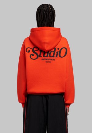 Personne aux cheveux tressés portant un sweat à capuche orange vif avec un texte noir "Studio Crafting Aesthetics WRSTBHVR" dans le dos, et un pantalon noir avec des bandes rouges.