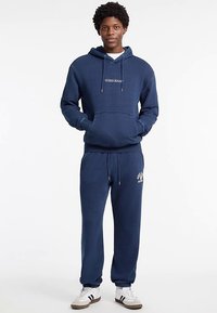 Sudadera azul marino de algodón con bolsillo canguro y cordones; combinada con pantalones jogger a juego que presentan un logo. El modelo lleva zapatillas blancas.