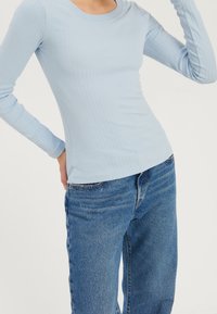 Kvinna iförd en ljusblå ribbad långärmad topp, instoppad i löst sittande blå jeans, stående mot en enfärgad bakgrund.