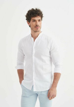 Chemise en lin blanche avec un col mao, manches longues retroussées au niveau des poignets et détails de boutons subtils, portée avec un pantalon bleu clair.