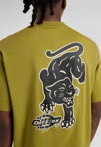 Modelka v zeleném tričku s velkým grafickým motivem černého pantera a stylizovaným textem "Dizzies" pod ním.