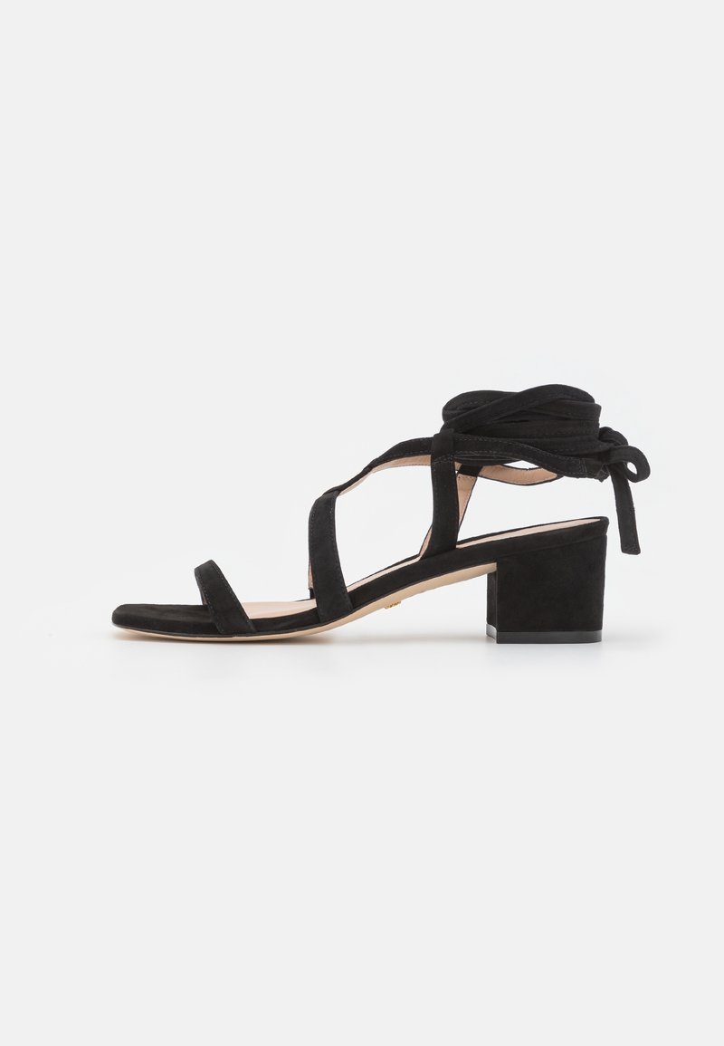 Sandale noire en daim avec lien à la cheville, bout ouvert, plusieurs brides et petit talon bloc sur fond blanc.
