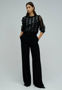 Haut noir transparent avec des appliqués floraux, manches longues, associé à un pantalon noir taille haute. Tissu doux, coupe ajustée, silhouette élégante.
