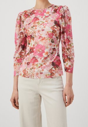 Woman wearing a long-sleeve, floral blouse with gathered shoulders and light-colored pants, standing against a plain background.