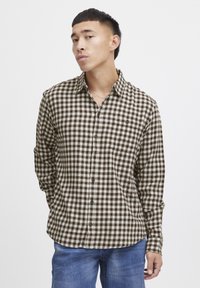 Camisa de franela a cuadros marrón y blanca con cuello, mangas largas, tapeta de botones y dobladillo redondeado. Combinada con jeans azules.