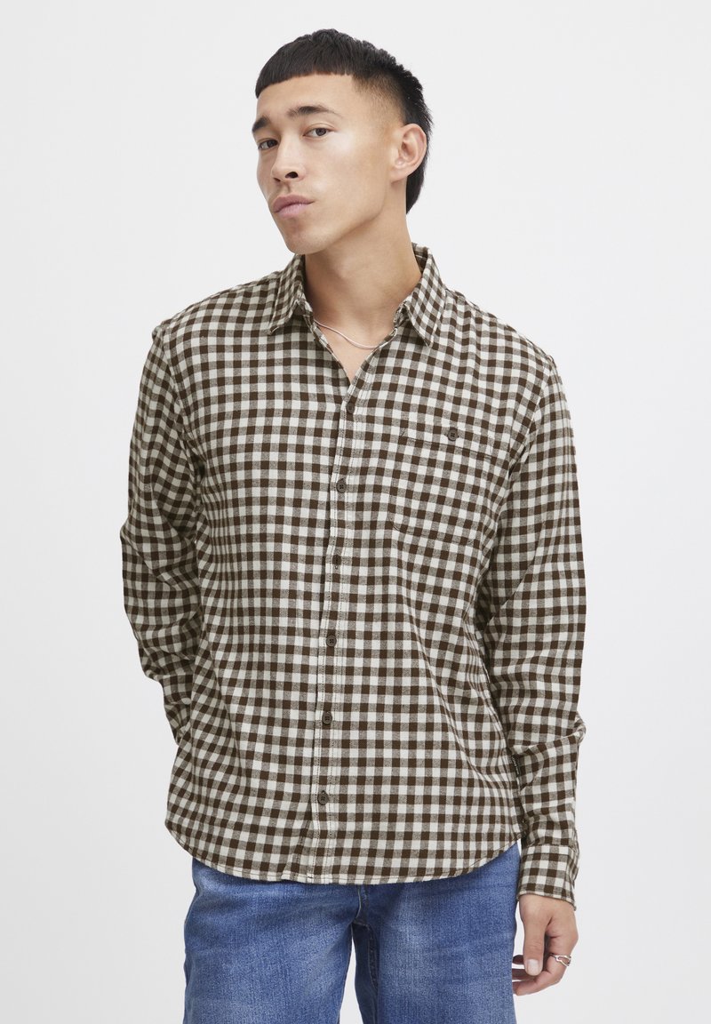 Camisa de franela a cuadros marrón y blanca con cuello, mangas largas, tapeta de botones y dobladillo redondeado. Combinada con jeans azules.