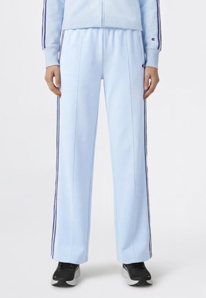 Jasnoniebieskie spodnie dresowe z ciemnymi bocznymi paskami, lekko rozszerzanymi nogawkami oraz czarno-szare buty sportowe, noszone przez stojącą osobę.