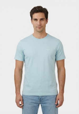 Homme portant un t-shirt bleu clair à manches courtes et un jean bleu, debout face à un fond blanc uni.