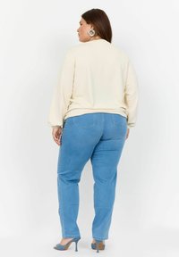 Cremefärgad långärmad topp med elastiska ärmslut, tillsammans med ljusblå raka jeans. Modellen bär grå klackskor och stora örhängen.
