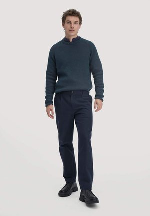 Dunkelblauer, gerippter Pullover mit hohem Kragen, kombiniert mit marineblauen Hosen und schwarzen, derben Stiefeln. Das Modell steht vor einem schlichten Hintergrund.
