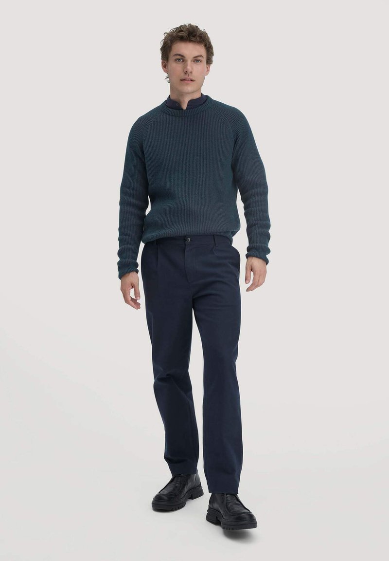 Donkerblauwe geribde trui met hoge kraag, gecombineerd met marineblauwe broek en zwarte chunkyschoenen. Model staat tegen een effen achtergrond.