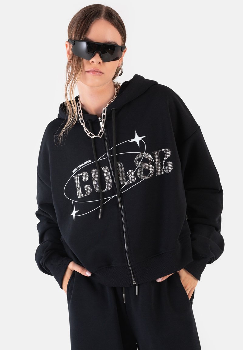 Czarna bluza z kapturem zapinana na zamek, wykonana z miękkiego materiału, ozdobiona logo i gwiazdkami z kryształków. Połączona z luźnymi czarnymi spodniami oraz masywnym srebrnym łańcuchem.