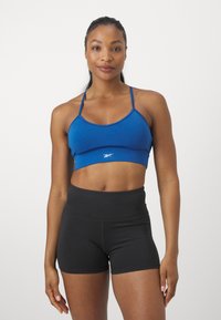 Μπρά με φωτεινό μπλε ύφασμα και λεπτές τιράντες, διακοσμημένο με λευκό λογότυπο, συνδυασμένο με μαύρες κοντές, ψηλόμεσες βερμούδες, και τα δυο φτιαγμένα για αθλητική χρήση.