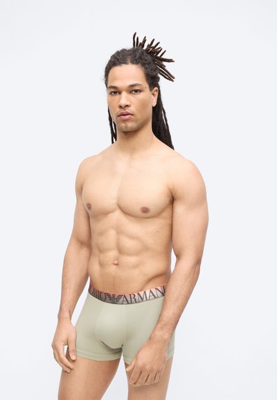 Μυώδης άνδρας με μακριές ντρέντλοκς που φοράει ανοιχτό πράσινο εσώρουχο Armani boxer στέκεται μπροστά σε απλό φόντο.