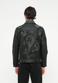 Jachetă din piele neagră cu mai multe patch-uri și studuri, având o textură netedă și un design casual, prezentând în mod proeminent diverse grafici pe spate.