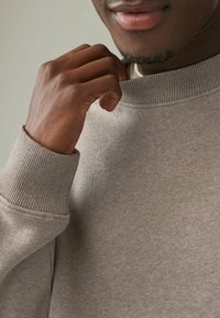 Felpa grigia in cotone con colletto e polsini a costine, caratterizzata da una texture liscia e un sottile motivo marmorizzato. Mano che tocca il colletto.