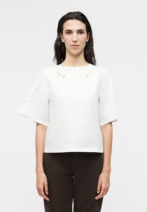 Γυναίκα με σκούρα μαλλιά που φοράει λευκό κοντομάνικο μπλουζάκι με μικρές λεπτομέρειες κοψίματος και μαύρο παντελόνι, κοιτάζοντας μπροστά σε απλό φόντο.