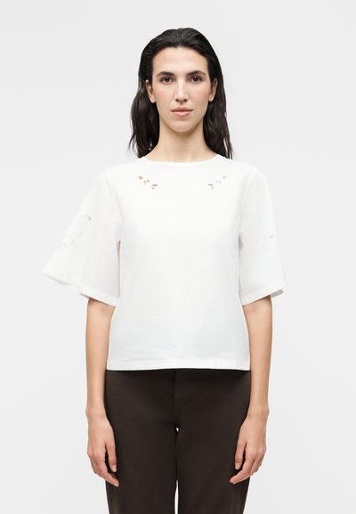 Femme aux cheveux foncés portant un haut blanc à manches courtes avec de petits détails découpés et un pantalon noir, face à un fond uni.
