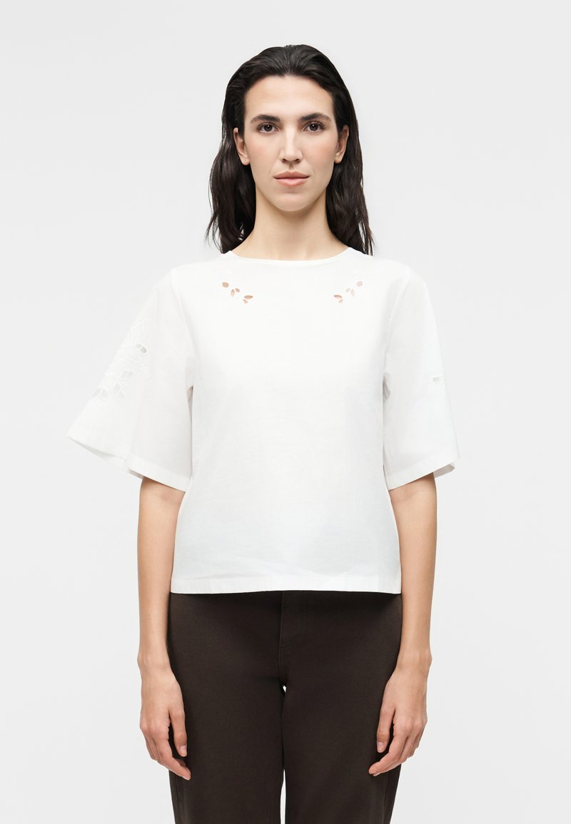 Γυναίκα με σκούρα μαλλιά που φοράει λευκό κοντομάνικο μπλουζάκι με μικρές λεπτομέρειες κοψίματος και μαύρο παντελόνι, κοιτάζοντας μπροστά σε απλό φόντο.