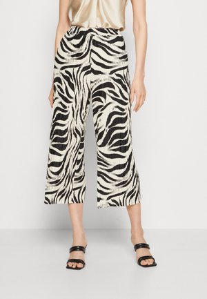 Kvinde iført plisserede, sort-hvide zebra-print bukser med vide ben og kort længde, sorte åbne hælesandaler og en satin beige top.