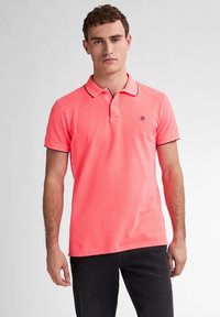 Chemise polo rose avec un col, des manches courtes et des bordures bleu marine le long du col et des manches. Logo sur la poitrine gauche. Texture en coton lisse.