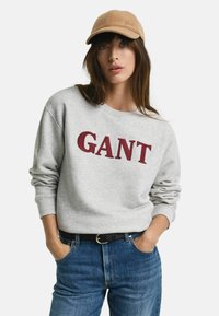 Grijze sweatshirt met rode "GANT" tekst, gedragen met blauwe spijkerbroek en een tan kleurige pet, met een zachte textuur en een relaxte pasvorm.