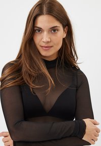 Sort gennemsigtig bluse med lange ærmer og høj hals, med et gennemsigtigt design og lagdelt stof over en mørk trekantet bh.
