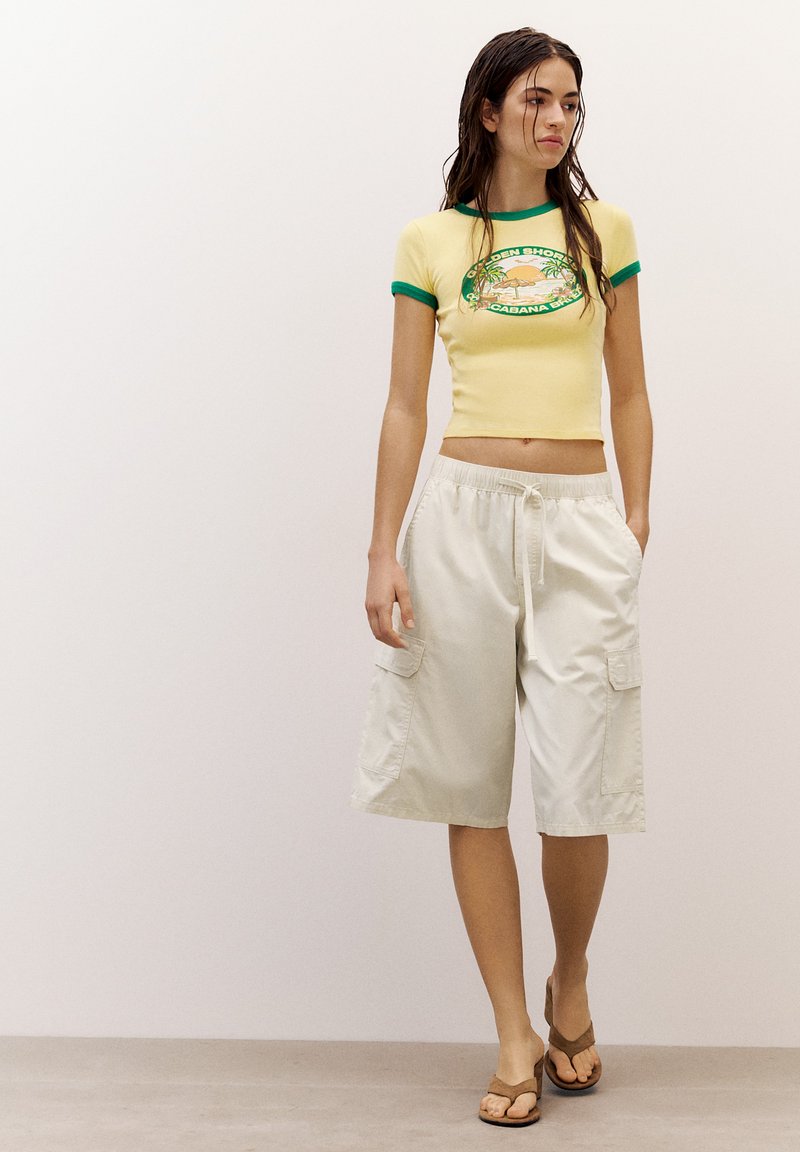 Young woman wearing a yellow crop top with green trim, beige long cargo shorts, and brown sandals standing against a plain background.