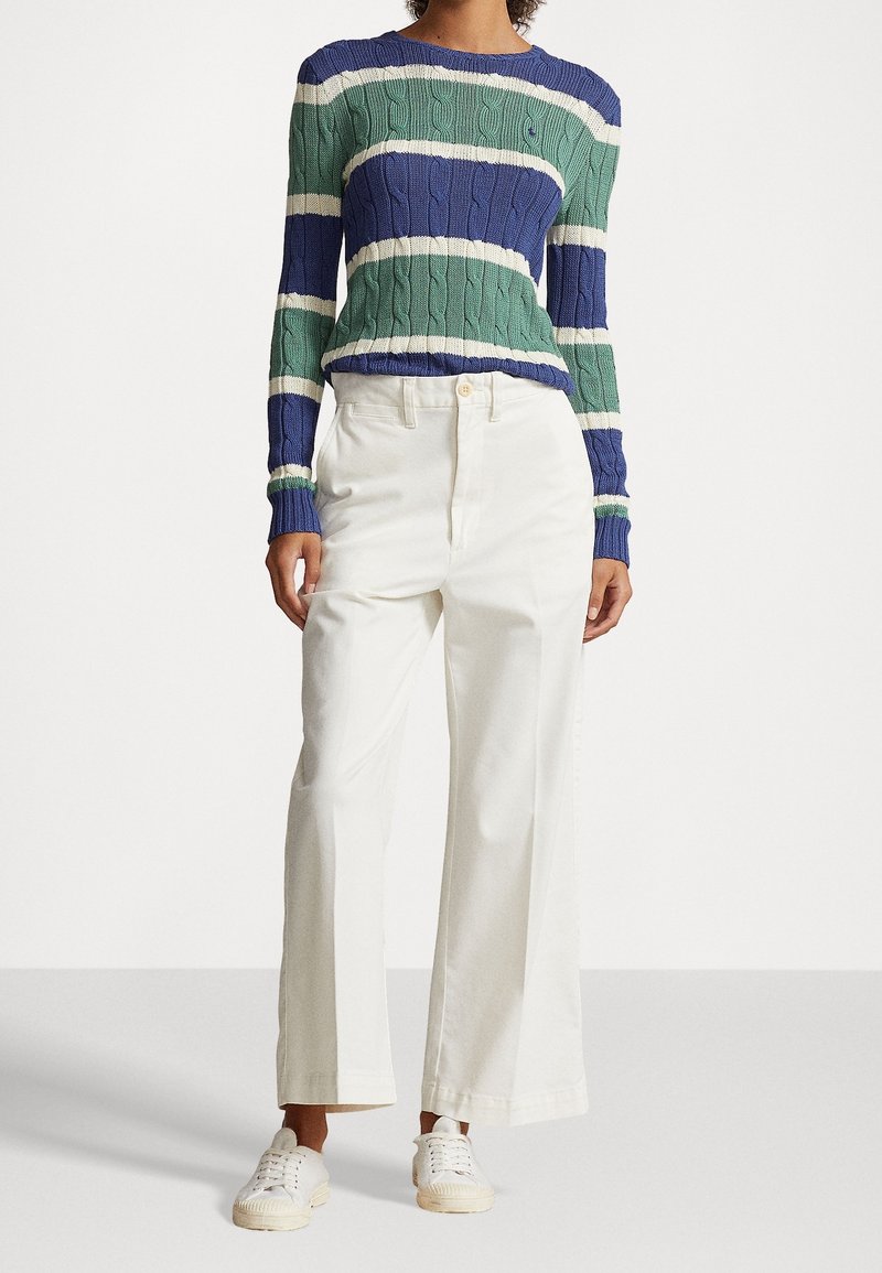 Mujer vestida con un suéter de punto trenzado a rayas azules y verdes, pantalones anchos de tiro alto blancos y zapatillas blancas, de pie sobre un fondo liso.
