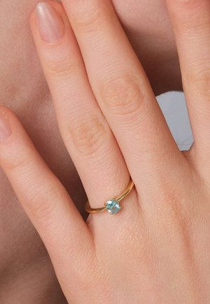 Left hand with a gold ring featuring a small oval blue gemstone worn on the ring finger against bare skin background.