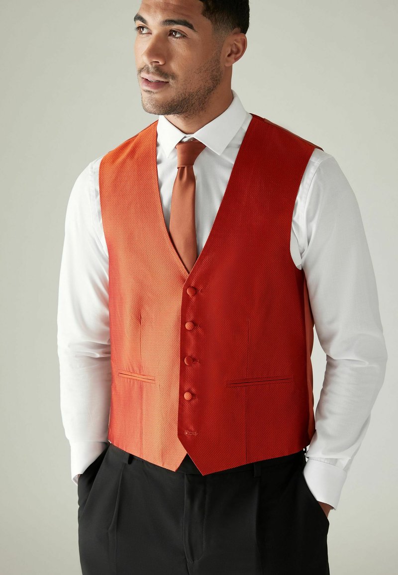 Gilet texturé rouge avec six boutons, associé à une cravate assortie et une chemise blanche. Design en V, forme ajustée et poches de chaque côté.